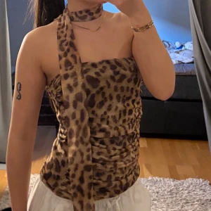 Leopardmönstrad topp med scarfdetalj -  leopardmönstrad topp med rak halsringning och en unik scarfdetalj som knyts runt halsen. Toppen är figurnära och har rynkade sömmar i sidan för en snygg passform. 