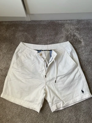 Vita shorts från Polo Ralph Lauren - Snygga vita shorts från Polo Ralph Lauren med klassisk blå logga broderad på benet. Shortsen har snörning i midjan, dragkedja och sidofickor. Perfekta för sommaren och tillverkade i mjuk bomull för skön känsla. Jag säljer dessa shortsen för att jag tyvärr växt ur dem. Storlek M