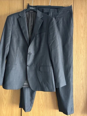 Oscar Jacobson tvådelad kostym - Säljer en väldigt fin kostym med både kavaj och byxor från Oscar Jacobson. Edmund suit.   I en väldigt snygg mörkare grå/brun färg. Finaste kvaliteten, tyg från Italien. Både kavaj och byxa är i storlek 50.   Endast använd ett fåtal gånger, jättebra skick.   Perfekt till bröllopet, studenten eller examen.