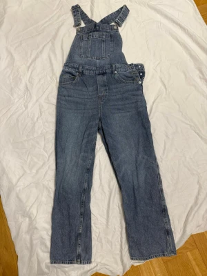 Klassisk blå jeansbyxdress - Säljer en klassisk blå jeansbyxdress med raka ben och justerbara hängslen. Byxdressen har en stor bröstficka, sidofickor och knappar i sidan för enkel påklädning. Perfekt för en avslappnad och trendig look. Materialet är slitstark denim i bomull.