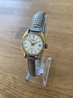 1981 Citizen Seven Date 24mm Damklocka - Ovanlig vintage Citizen Seven damklocka med datumvisning. Klockan har ett guldpläterat boett och ett tvåfärgat (silver och guld) flexibelt armband. Baksidan av boetten är märkt med "Citizen WATCH CO. BASE METAL TOP ST. STEEL BACK 4-014170 SMN 1090340 BV-2065 GP". Armbandet är inte original men högkvalitativt märkt "Timex Stainless Steel". Japanskt urverk. Fint använt skick enligt bilder. Behöver bara nytt batteri.  Värde: 900 SEK Mitt pris: 589 SEK