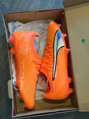 Puma Ultra orange fotbollsskor - Snygga och lätta Puma Ultra fotbollsskor i en riktigt skarp orange färg med svart och vit detalj på sidan. Skorna har låg profil, snörning och en ovandel i syntetmaterial som ger bra bollkontroll och grepp på planen. Perfekta för dig som vill sticka ut på matchen.