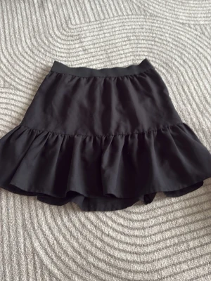 Svart volangskort från Gina Tricot - Supersöt svart skort från Gina Tricot med bred volangkant och elastisk midja. Kjol framtill och shorts under, perfekt för en trendig och bekväm look. Tillverkad i ett mjukt, lätt tyg som ger en snygg siluett.