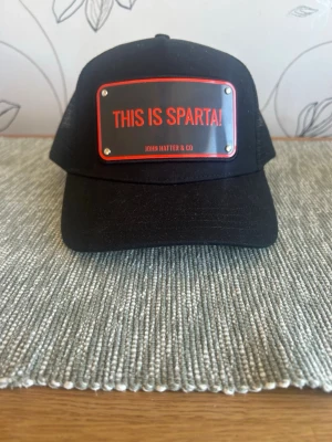 Svart keps John Hatter & Co - Snygg helt oanvänd svart keps från John Hatter & Co med en metallskylt framtill där det står 'THIS IS SPARTA!'. Kepsen har böjd skärm och mesh på sidorna. Som sagt helt oanvänd och mycket bra skick, 380 kr