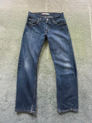 Levi's 512 Bootcut jeans blå - Säljer ett par klassiska Levi's 512 Bootcut jeans i blå denim. Jeansen har fem fickor, orange sömmar och en lätt utsvängd passform vid benslutet. Perfekta för dig som gillar en tidlös och avslappnad stil. Mycket gott skick, som syns på bilderna, med synliga lagningar. Storlek: W30 L32 Ungefärliga mått: Midjevidd - 38 cm Lårvidd - 27 cm Längd från midja - 98,5 cm Längd från grenen - 74,5 cm Innersöm - 20 cm