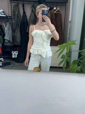 Vit volang topp med smala axelband - Säljer en superfin vit topp med smala axelband och volangdetaljer. Toppen har en ribbad struktur och är figurnära med en snygg omlottkänsla framtill. Perfekt för dig som vill ha något unikt och trendigt i garderoben!