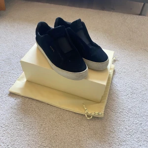 Svarta sneakers från Axel Arigato - Stilrena svarta sneakers från Axel Arigato i mocka och skinn med vit platt sula. Diskret guldtext på sidan och minimalistisk design utan synlig snörning. Perfekta för dig som gillar en clean och modern look.