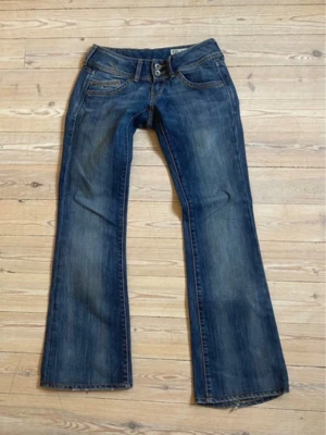 Blå Lågmidjade bootcut jeans med dubbla knappar - Snygga blå bootcut jeans med dubbla knappar i midjan och klassiska fem fickor. Jeansen har en lätt tvättad look och subtila slitningar för en cool stil. Perfekta för dig som gillar en avslappnad men trendig vibe.