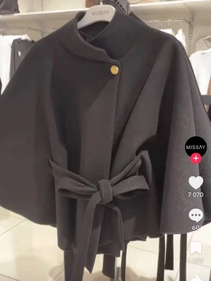 Svart cape-kappa från Missay - Stilren svart cape-kappa från Missay med knytband i midjan och en dekorativ guldfärgad knapp vid halsen. Kappan har en vid, svepande siluett och är tillverkad i ett mjukt tyg som ger en elegant look. Perfekt för dig som vill ha en trendig och unik ytterjacka.