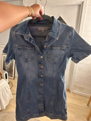 Jeansklänning från ZARA med knappar - Snygg jeansklänning från ZARA i blå denim med kort ärm och knappar framtill. Klänningen har två bröstfickor och klassisk krage, perfekt för en trendig och avslappnad look. Storleken är L men passar mycket bra en M