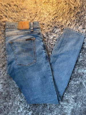 Ljusblå skinny jeans från Nudie - Säljer ett par ljusblå skinny jeans från Nudie med klassisk femficksdesign och snygga detaljer på bakfickan. Jeansen har en smal passform och är tillverkade i mjuk denim med stretch för extra komfort.