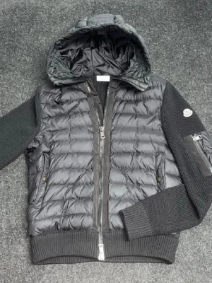 Svart pufferjacka från Moncler - Snygg svart pufferjacka från Moncler med quiltad front och huva. Ärmar och rygg är i stickad ullmix, vilket ger en cool kontrast. Jackan har dragkedja fram, fickor med tryckknapp och en Moncler-logga på ärmen. Perfekt för kalla vinterdagar.