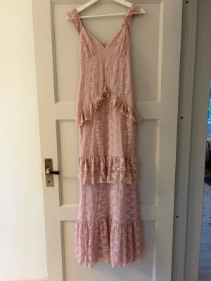 Ljusrosa spetsklänning från Nelly - Gammelrosa långklänning i spets med volangdetaljer och snörning i ryggen. Klänningen har axelband och v-ringning, samt flera lager volanger längs kjolen. Perfekt nu inför balen eller andra stora fester. Klänningen är oanvänd och har inga defekter. Prislappen sitter kvar.