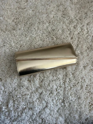 Guldig kuvertväska med kedja - Snygg kuvertväska i metallic guld med stilren design och magnetlås från Anna Field. Väskan har en avtagbar tunn kedja i guld som gör att den kan bäras både i handen och över axeln. Tillverkad i glansigt konstläder. Använd en gång, så som ny