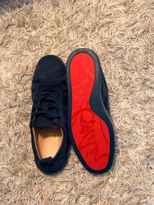 Mörkblå Christian Louboutin sneakers - Snygga mörkblå sneakers från Christian Louboutin med klassisk röd sula och diskret logga. Skorna är låga och har ovandel i mocka med matchande mörkblå snörning. Perfekta för dig som vill sticka ut med exklusiva detaljer och stilren design.