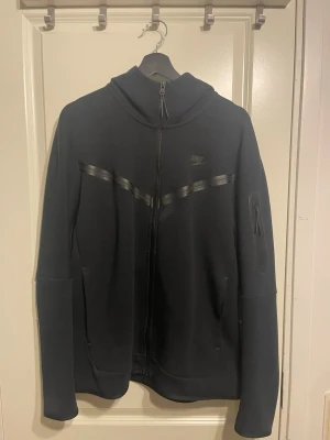 Svart Nike hoodie med dragkedja - Snygg svart hoodie från Nike med hel dragkedja framtill och diskret Nike-logga på bröstet. Hoodien har en huva, två sidofickor och en extra ficka med dragkedja på ärmen. Materialet är mjukt och stretchigt, perfekt för en avslappnad stil.