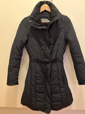 Svart quiltad rock från Calvin Klein - Lång svart quiltad rock från Calvin Klein Jeans med hög krage och bälte i midjan. Rocken har dragkedja och tryckknappar framtill samt ett justerbart bälte med logotyp. Tillverkad i ett lätt, vadderat material som håller värmen.