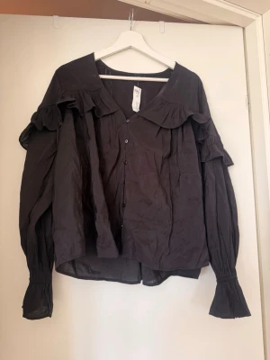 Svart volangblus med knappar - Snygg svart blus från Lindex med volangdetaljer längs axlarna och knäppning framtill. Blusen har långa ärmar med breda, rynkade muddar och en lös passform. Perfekt för dig som gillar trendiga detaljer och en chic look.