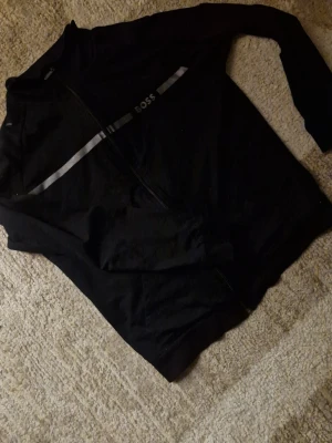 Svart zip-crewneck från BOSS - Säljer en svart crewneck från BOSS med hel dragkedja och grå detaljer på bröstet. Hoodien har ett stilrent och sportigt utseende, tillverkad i mjukt material som känns skönt mot huden. Perfekt för dig som gillar klassiska märken och enkel design. Passformen är stilren, då den sitter utmärkt för nästan vem som helst. En fin boss tröja i utmärkt sick, det kan inte gå fel. Fast pris, ett mycket rimligt och inte förhandlingsbordet pris. Kom pv för mer info!