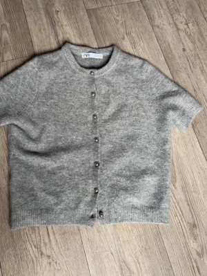 Grå kortärmad kofta från Zara - Mysig grå kofta från Zara med korta ärmar och rund hals. Koftan har knappar framtill i metall och är stickad i ett mjukt material. Knapparna går dessutom att öppna!!💕💕💕(skulle säga att den är liten i storlek)