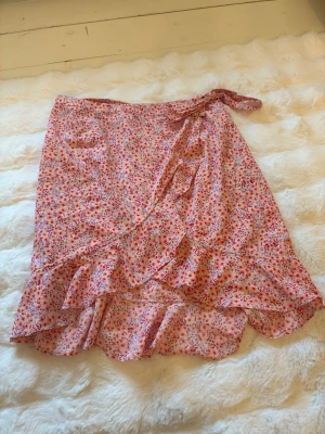 Blommig omlottkjol med volang - Söt omlottkjol med blommigt mönster i rosa, rött och gult. Kjolen har volangkant och knyts i midjan för en feminin look. Perfekt för varma dagar och ger en lätt och luftig känsla.