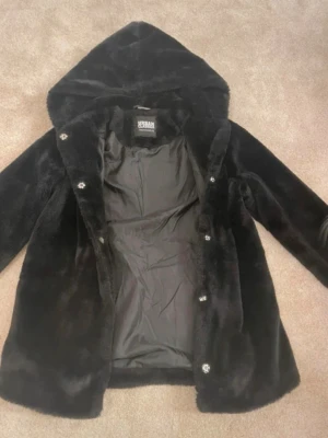 Svart fuskpälsjacka med huva Urban - Snygg svart fuskpälsjacka från Urban Classics med huva och knäppning framtill. Jackan har en mjuk och fluffig yta, helsvart innerfoder och raka ärmar. Perfekt för dig som vill ha en varm och trendig vinterjacka med lite extra attityd.