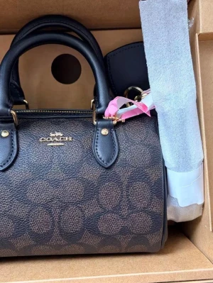 Brun monogram handväska från Coach - Snygg brun handväska från Coach med klassiskt monogrammönster och svarta handtag i läder. Väskan har guldfärgade detaljer och dragkedja upptill. Perfekt accessoar för dig som gillar stilrena och trendiga väskor.