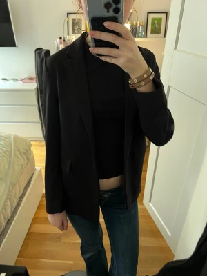 Svart klassisk kavaj med smal krage - Stilren svart kavaj med smal krage och enkelknäppning. Modellen har två fickor framtill och en figurnära passform som ger en snygg siluett. Perfekt att styla med jeans eller dressade byxor. Materialet ser ut att vara en mjuk och slät textil. Barnstorlek men skulle säga att den passar som xs 