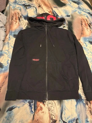 Svart hoodie från Hugo med dragkedja - Svart hoodie från Hugo med stor röd logga på huvan och mindre logga på bröstet. Hoodien har hel dragkedja, snörning i huvan och känguruficka framtill. Tillverkad i mjuk bomull, perfekt för en chill och stilren look.