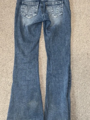 Blå bootcut jeans med slitningar - Säljer ett par blå bootcut jeans med snygga slitningar och klassiska bakfickor med V-detalj. Jeansen har en lätt utsvängd passform och är perfekta för en avslappnad och trendig stil. Tillverkade i bomull för skön känsla.