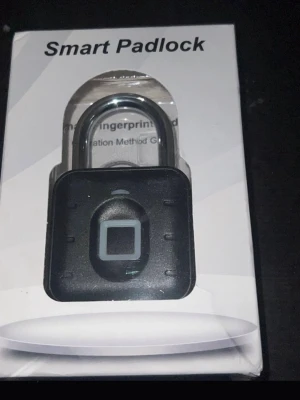 Smart hänglås med fingeravtrycksläsare - Elektroniskt smart hänglås i svart med fingeravtrycksläsare på framsidan. Låset har en fyrkantig form och är tillverkat i metall och plast. Perfekt för att låsa in dina prylar på ett smidigt och modernt sätt.