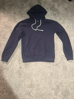 Mörkblå hoodie pier one  - En stilren mörkblå hoodie med huva och vita dragsnören. Hoodien har en klassisk känguruficka framtill och ribbade muddar vid ärmslut och nederkant. Perfekt för en avslappnad look. Materialet känns mjukt och bekvämt. Storlek S men liten i storlek så kan även passa XS.