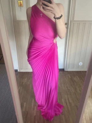 Rosa plisserad långklänning oneshoulder - Snygg rosa långklänning med plisserat tyg och oneshoulder-design. Klänningen har en elegant drapering och en liten cut-out vid midjan som ger en modern touch. Perfekt för dig som vill sticka ut med en färgstark och trendig look. Använt 1 gång som ny!! Passar även S