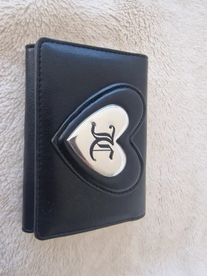 Svart plånbok med hjärtdekor Juicy Couture  - Snygg svart plånbok från Juicy Couture i konstläder med ett stort hjärta i silvermetall på framsidan och JC-logga. Plånboken har en stilren och ungdomlig design, perfekt för dig som gillar detaljer och vill ha något unikt i väskan.
