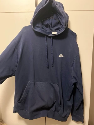 Blå Nike hoodie med huva - Mörkblå hoodie från Nike med klassisk logga på bröstet. Tröjan har stor huva med dragsko och en rymlig magficka. Tillverkad i mjukt bomullsmaterial och har en avslappnad passform, perfekt för en chill och sportig stil.