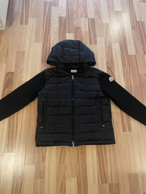 Svart Moncler cardigan - Svart cardigan från Moncler med huva och dragkedja framtill. Jackan har quiltad front i glansigt material, släta svarta ärmar och två fickor med dragkedja. Klassisk Moncler-logga på ena ärmen och randigt band som skickas med. Den var för liten för mig så den har aldrig kommit t användning. Kan släppa den billigare vid snabb affär