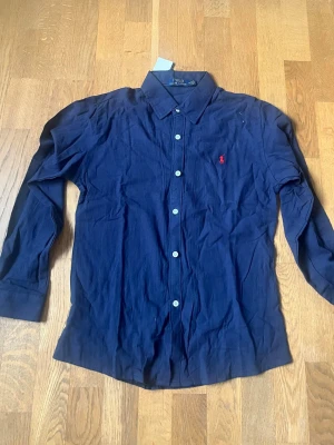 Marinblå skjorta från Polo Ralph Lauren - Säljer en klassisk marinblå skjorta från Polo Ralph Lauren med vit knappstängning och ikonisk röd logobrodyr på bröstet. Skjortan har lång ärm och är tillverkad i bomull, perfekt för en stilren look.