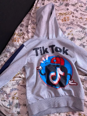 Grå hoodie med TikTok-tryck - Grå hoodie med stor färgglad TikTok-grafik på ryggen och texten 'TikTok'. Har huva och mörkblå rand längs ärmarna. Mjuk och bekväm, perfekt för dig som gillar streetstyle och sociala medier. Materialet känns som bomullsblandning.