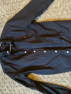 Mörkblå skjorta från Ralph Lauren - Klassisk mörkblå skjorta från Ralph Lauren med vit knappstängning och röd broderad logga på bröstet. Skjortan har lång ärm och är tillverkad i bomull, perfekt för en stilren look.