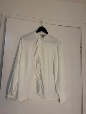 Vit blus med volang framtill - Stilren vit blus med lång ärm och volangdetalj längs knäppningen framtill. Blusen har klassisk krage och breda manschetter. Perfekt för en trendig och chic look. Materialet känns lätt och luftigt.