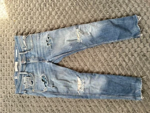 Ljusblå slitna jeans från Replay - Säljer ett par ljusblå jeans från Replay, modell Anbass. Jeansen har flera slitningar och hål för en trendig, avslappnad look. Klassisk femficksmodell med dragkedjegylf och raka ben. Perfekta för dig som gillar streetstyle.