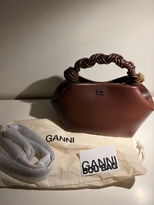 Brun handväska från Ganni  - Snygg brun handväska från Ganni med unik flätad handtag i samma färg. Väskan har en geometrisk form och är tillverkad i slätt läder. Inuti finns ett fack med Ganni-logga. Kommer med dustbag och extra axelrem. Ganni bou bag small