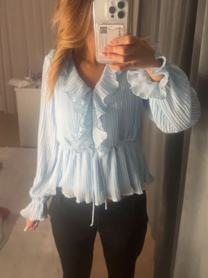 Ljusblå plisserad blus med volanger - Supersöt ljusblå blus med plisserat tyg och volangdetaljer längs v-ringningen och ärmsluten. Blusen har långärmad design och markerad midja som ger en fin siluett. Perfekt för dig som gillar romantisk och trendig stil.💙