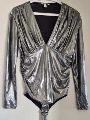 Metallic silver body med djup v-ringning - Snygg body i metallic silver med lång ärm och djup v-ringning. Materialet är glansigt och stretchigt, vilket ger en cool och trendig look. Dragsydda detaljer framtill och klassisk bodyform med knäppning nedtill. Så cool till årets alla fester!