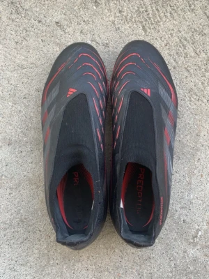 Adidas Predator svarta fotbollsskor - Säljer ett par svarta Adidas Predator fotbollsskor med röda och grå detaljer. Skorna har en strumpliknande passform utan snörning och mönstrad ovandel för extra grepp på bollen. Yttersulan har fasta dobbar för spel på gräsplan. Perfekta för dig som vill ha kontroll och stil på planen.