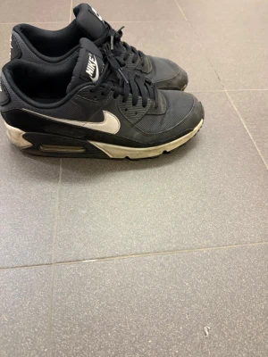 Nike Air Max 90 sneakers - Svarta Nike Air Max sneakers med vit sula och vit swoosh på sidan. Skorna har klassisk låg profil, mesh och syntetmaterial i ovandelen samt synlig Air Max-dämpning i hälen. Perfekta för en sportig och streetinspirerad stil.