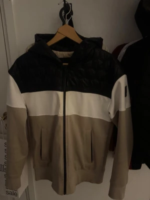 Beige och svart jacka från Moose Knuckles - Snygg jacka från Moose Knuckles med huva och dragkedja. Jackan har färgblock i svart, vitt och beige samt quiltad ovandel. Märkets logga sitter på ärmen. Perfekt för dig som gillar streetwear och stilren design.