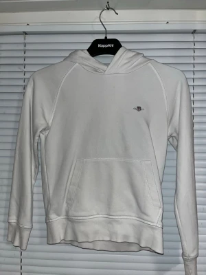 Vit hoodie från GANT med logga - Snygg vit hoodie från GANT med liten broderad logga på bröstet. Hoodien har klassisk känguruficka framtill. Väldigt fräsch tröja som passar till allt. 