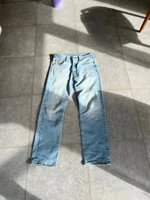 Levis 501 W31 L32 - Säljer ett par klassiska ljusblå Levi's jeans med rak passform och fem fickor. Jeansen har en vintage look med slitningar och är tillverkade i 100% bomull. Perfekta för en avslappnad och trendig stil.