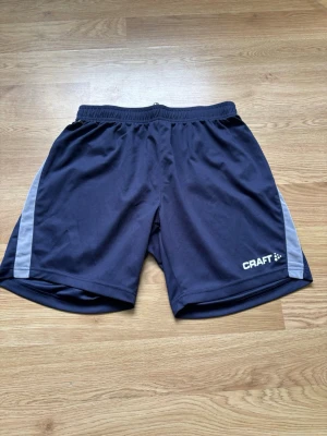 Svarta träningsshorts från Craft - Svarta träningsshorts från Craft med gråa sidopaneler och elastisk midja. Perfekta för sport eller gym. Lätta och bekväma i syntetmaterial som andas. Klassisk sportig look med logga på vänster ben.